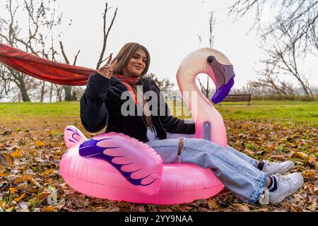 giovane bella donna che si diverte a cavalcare un fenicottero rosa gonfiabile in un parco pubblico in inverno Foto Stock