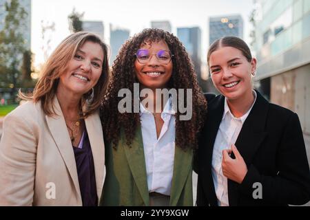Tre donne d'affari fiduciose sorridono guardando la macchina fotografica. Lavoro di squadra, diversità, professionalità e successo mentre si trovano insieme all'aperto, irradiando, felicità ed energia positiva sul posto di lavoro. Foto di alta qualità Foto Stock
