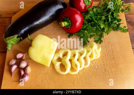 Il peperone giallo appena tritato forma spirali su un tagliere di legno. Nelle vicinanze ci sono melanzane, peperoni rossi, chiodi di garofano e rametti di parsle Foto Stock