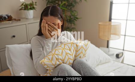 Giovane donna seduta nella sua camera da letto che ricopre il viso con le mani tenendo un cuscino decorativo, circondata da un arredamento accogliente e da un'illuminazione soffusa Foto Stock