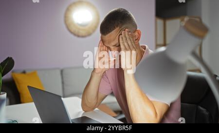 Giovane con mal di testa seduto in un salotto di casa, tenendo la testa con entrambe le mani, apparendo stressato e frustrato mentre guarda un grembo Foto Stock