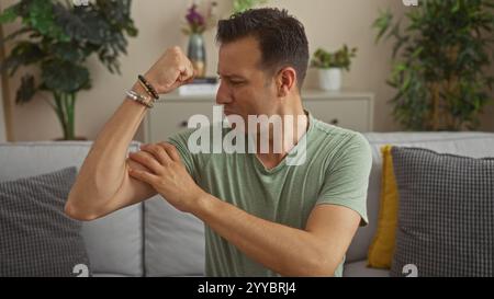 Uomo ispanico con una camicia verde che si flette i muscoli mentre si siede in un accogliente soggiorno con piante e cuscini intorno, mostrando forza in casa. Foto Stock
