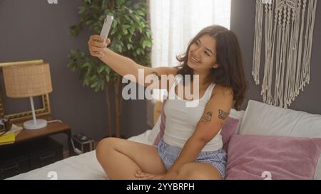 Una giovane donna scatta un selfie mentre si siede su un letto in una camera da letto accogliente, mostrando i suoi capelli bruna e i suoi tatuaggi in un ambiente domestico. Foto Stock