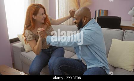 Coppia interrazziale che ride insieme in un accogliente soggiorno, mostrando amore e felicità al chiuso con un uomo e una donna che si godono una calda e intima momen Foto Stock