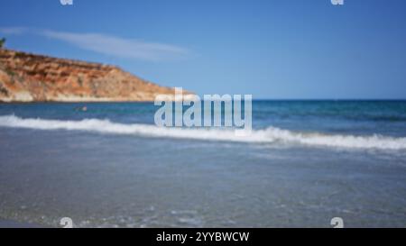 Vista sfocata di una splendida spiaggia con onde che si infrangono contro la riva, circondata da cielo azzurro e scogliere rocciose sullo sfondo Foto Stock