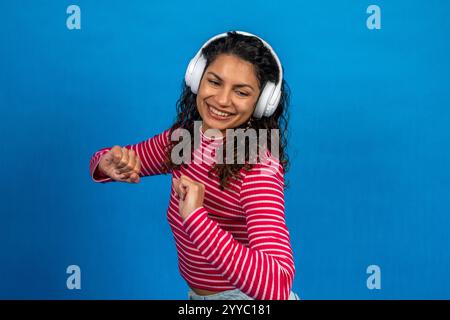 Giovane donna spensierata che ascolta la musica, indossa le cuffie wireless e balla con gioia su uno sfondo blu brillante Foto Stock