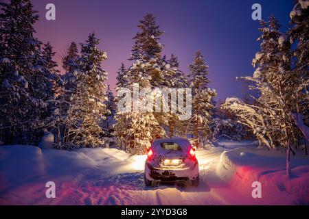 Lapponia finlandese in inverno. Foresta innevata di notte. Auto innevata con fari sulla strada forestale Foto Stock