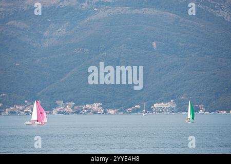24 ottobre 2024, Tivat, Montenegro: Due barche a vela che navigano in mare al tramonto con montagne e una città costiera sullo sfondo Foto Stock