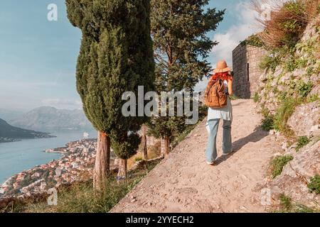 Donna turistica che indossa cappello e zaino camminando lungo il sentiero sterrato lungo il muro di pietra che si affaccia sulla baia di Cattaro in Montenegro Foto Stock