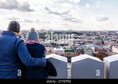 Una coppia sta in piedi su una sporgenza che si affaccia su una città. L'uomo indossa una giacca blu e la donna indossa una sciarpa rosa. Il cielo è nuvoloso e.. Foto Stock