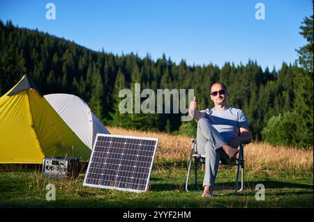 Uomo che utilizza un pannello solare fotovoltaico per caricare una centrale elettrica portatile all'aperto in campeggio ecologico. Un turista di sesso maschile che dà i pollici vicino alle tende. Sullo sfondo alti pini e cielo azzurro. Foto Stock