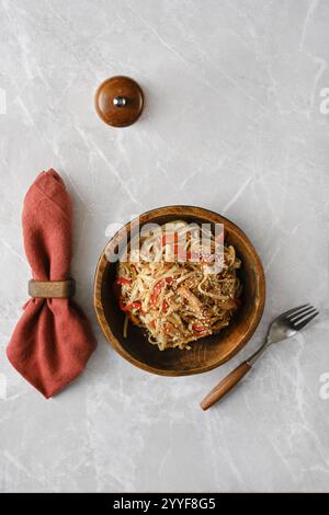 Una ciotola di legno ripiena di spaghetti saltati mescolati con manzo tirato, peperoni e condita con semi di sesamo poggia su un tavolo di marmo, accompagnato da Foto Stock