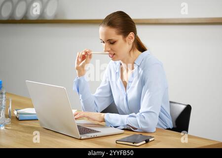 Una donna è profondamente impegnata mentre usa il suo notebook in un ufficio contemporaneo. Si siede su un tavolo di legno, circondato da un ambiente tranquillo, indicando pr Foto Stock