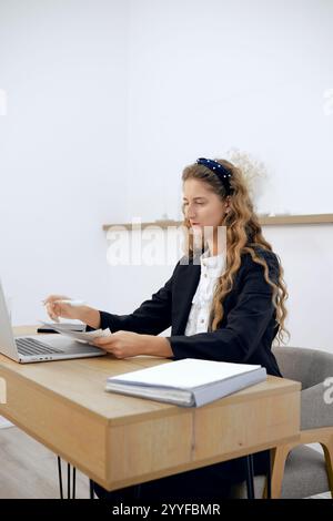 Una giovane donna concentrata con capelli ricci siede su una scrivania minimalista, rivede i documenti e scrive sul suo notebook. La camera luminosa e dai colori neutri è vuota Foto Stock