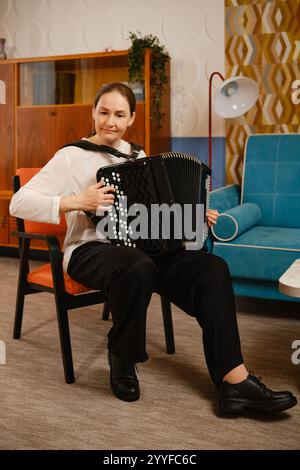 Una donna di mezza età è seduta comodamente in un salotto vintage, suonando sapientemente la fisarmonica. Foto Stock