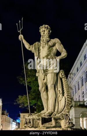 Una statua di un uomo che regge un tridente è in piedi di fronte a un edificio. La statua è fatta di pietra ed è illuminata dai lampioni Foto Stock