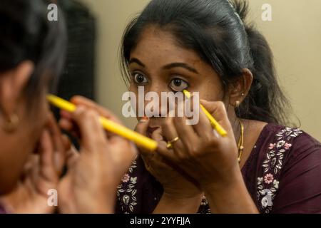 Una giovane donna dell'Asia meridionale applica con attenzione l'eyeliner, riflettendo nello specchio. Foto Stock