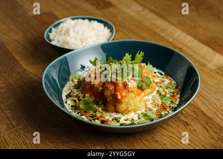 Un piatto a base di cavolfiore arrosto condito con una salsa piccante, guarnito con erbe fresche e servito in una ciotola blu. Una ciotola di accompagnamento di riso al vapore Foto Stock