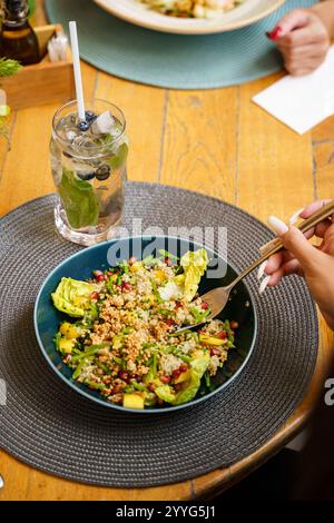 Un'insalata colorata di quinoa con verdure fresche, semi di melograno e verdure tritate servite in una ciotola blu. Una bevanda rinfrescante con ghiaccio, menta e blu Foto Stock