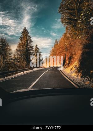 Una vista pittoresca dall'interno di un'auto su una strada boschiva tortuosa circondata da alberi spolverati di neve e illuminata dalla calda luce del sole. Foto Stock