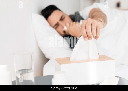 Giovane malato sdraiato a letto e starnuto che cola, prendendo i tovaglioli, concentrandosi selettivamente sulla scatola dei tessuti sul comodino Foto Stock