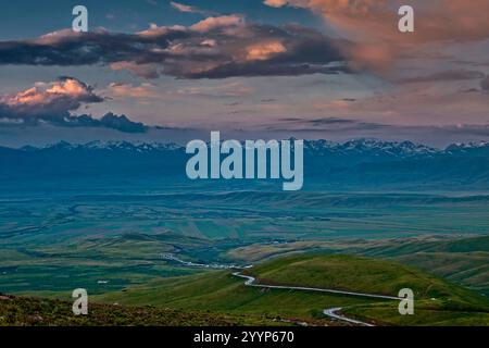 La luce del mattino illumina la valle di Suusamyr in Kirghizistan. Una strada tortuosa conduce attraverso verdi colline verso montagne innevate. Foto Stock