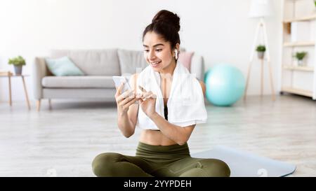 Affascinante giovane signora indiana con auricolari e telefono cellulare scelta playlist musicale per l'allenamento domestico Foto Stock