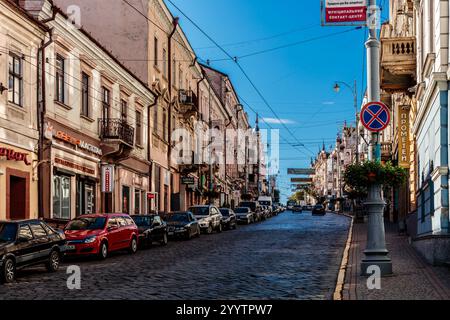 Una strada con un'auto rossa parcheggiata sul lato della strada. La strada è costeggiata da edifici e ci sono molte auto parcheggiate Foto Stock