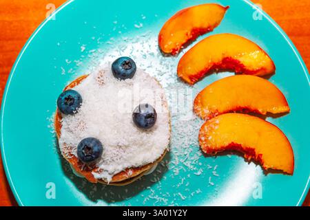 Pancake impilati con scaglie di cocco e mirtilli, accompagnati da fette di pesca. Pancake appena fatti, adornati con scaglie di cocco Foto Stock