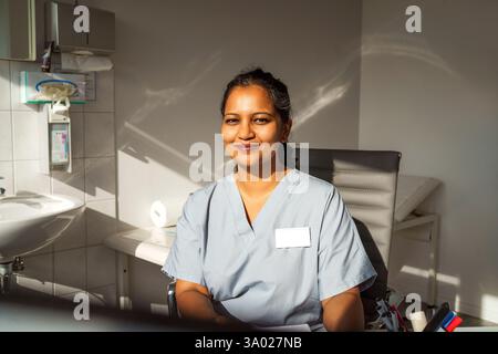 Ritratto di un medico sorridente che indossa scrub seduto sulla sedia nello studio del medico all'ospedale Foto Stock