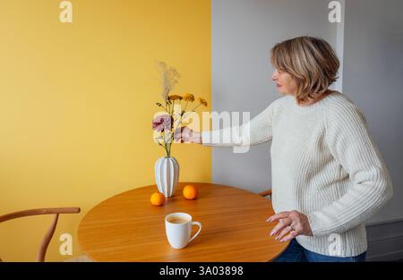 Una donna anziana con un maglione bianco e un jeans raccoglie un bouquet autunnale in un vaso su un tavolo rotondo in legno. Una donna matura decora la sua casa con flo Foto Stock