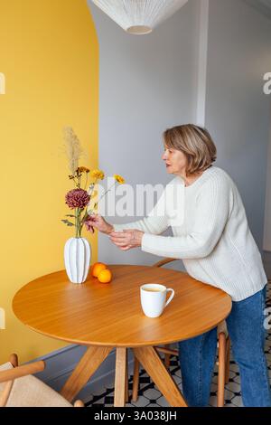 Una donna anziana con un maglione bianco e un jeans raccoglie un bouquet autunnale in un vaso su un tavolo rotondo in legno. Una donna matura decora la sua casa con flo Foto Stock