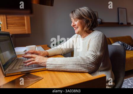 Una donna anziana in abiti casual sta lavorando su un notebook. La signora matura che ride sta scrivendo su un computer. Il pensionato sorride e guarda il grembo Foto Stock