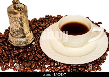 Macinacaffè vintage con design in metallo ornato e tazza da caffè bianca classica su un piattino, circondato da chicchi di caffè torrefatti Foto Stock