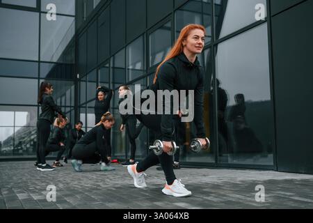Sollevamento manubri. Un gruppo di donne sportive si trova all'aperto vicino all'edificio nero. Foto Stock