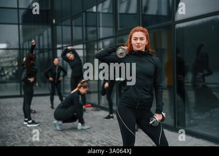 Sollevamento manubri. Un gruppo di donne sportive si trova all'aperto vicino all'edificio nero. Foto Stock