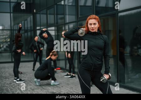 Sollevamento manubri. Un gruppo di donne sportive si trova all'aperto vicino all'edificio nero. Foto Stock