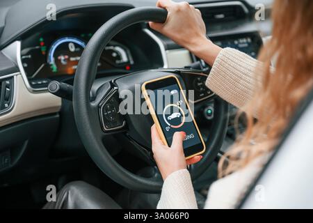 Seduto al volante e tenendo in mano lo smartphone che mostra il processo di carica. La giovane donna è nell'automobile. Foto Stock