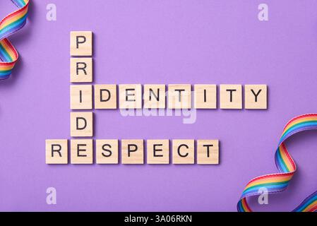 Pezzi di legno con lettere che formano un puzzle di parole incrociate con le parole orgoglio, identità, rispetto, accanto ai nastri della bandiera arcobaleno, su sfondo viola. Po Foto Stock