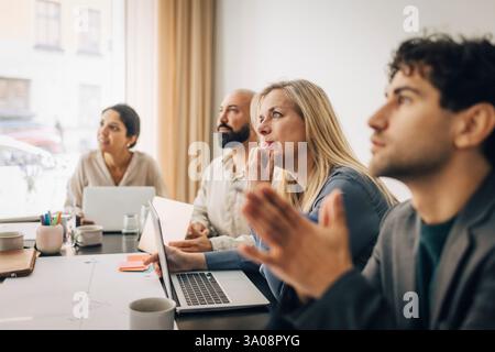 Donna d'affari matura concentrata seduta con un computer portatile al tavolo vicino a colleghi di sesso maschile durante la riunione in ufficio Foto Stock