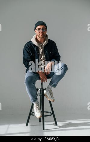 Il modello in cappello e occhiali è seduto su una sedia. Bell'uomo e' nello studio su sfondo bianco. Foto Stock
