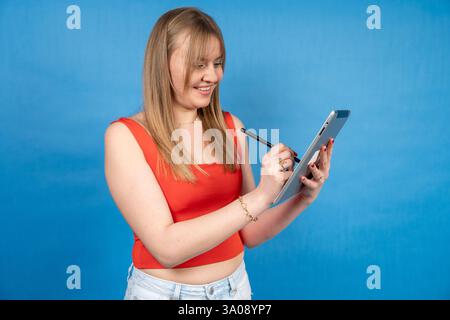 Giovane donna sorridente che utilizza la penna stilo per disegnare su un tablet digitale, godendo di un processo creativo su uno sfondo blu brillante Foto Stock