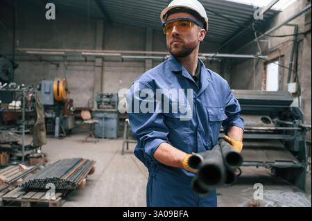 Guardando lateralmente e tenendo i tubi metallici. Il lavoratore di fabbrica in uniforme blu è al chiuso. Foto Stock