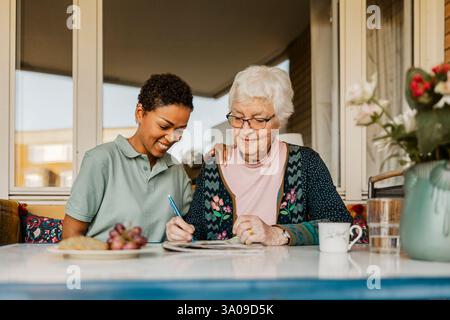 Donna anziana che risolve un puzzle di parole incrociate sul giornale mentre siede accanto a un'infermiera donna fuori casa Foto Stock