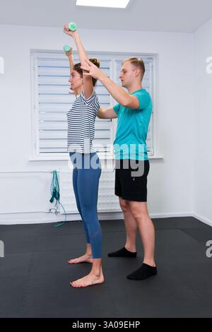 Donna con dolore alla spalla in terapia fisica. Fisioterapista che si esercita con una paziente che fa esercizi di mobilità delle spalle utilizzando un leggero Foto Stock