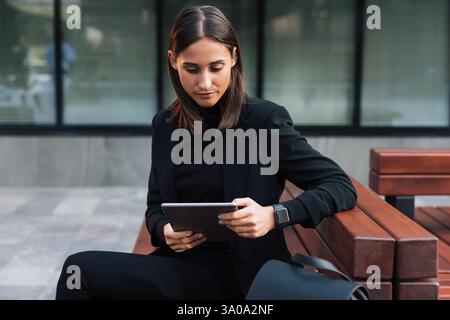 Giovane donna con un tablet digitale seduto all'aperto che indossa un elegante abbigliamento formale nero. Donna d'affari che legge da un tablet digitale. Foto Stock