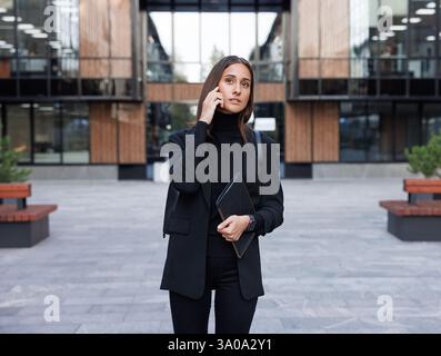 Giovane imprenditrice che indossa abiti eleganti e formali neri con colletto alto che effettua una telefonata nel cortile di campagna di un edificio per uffici Foto Stock