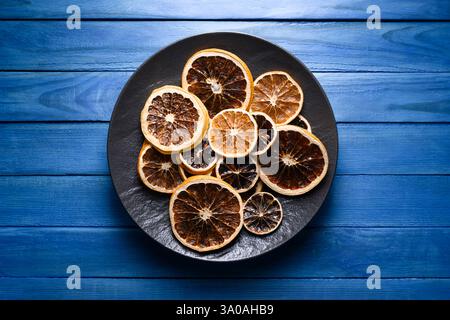 Fette di agrumi essiccate in piatto nero su un tavolo rustico in legno blu. Fotografia gastronomica Foto Stock