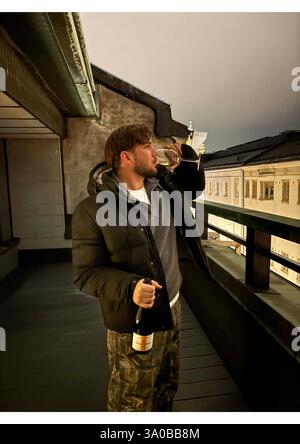 'Un uomo elegante si gode un bicchiere di champagne su un balcone sul tetto di Helsinki, in Finlandia, con lo skyline della città illuminato sullo sfondo. Vestito Foto Stock