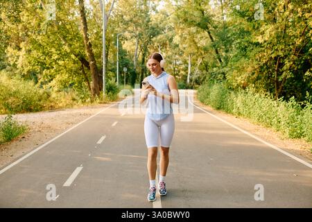 Una giovane atleta si prende una pausa su un sentiero boscoso, controllando il telefono con un sorriso. Foto Stock